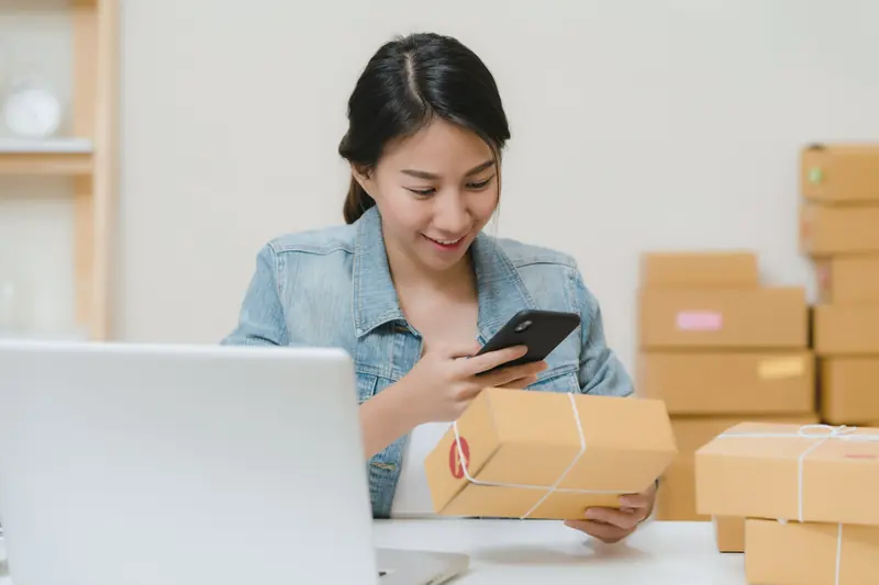 Người phụ nữ dùng điện thoại kiểm tra một kiện hàng, bên cạnh máy tính xách tay và các thùng hàng xếp chồng